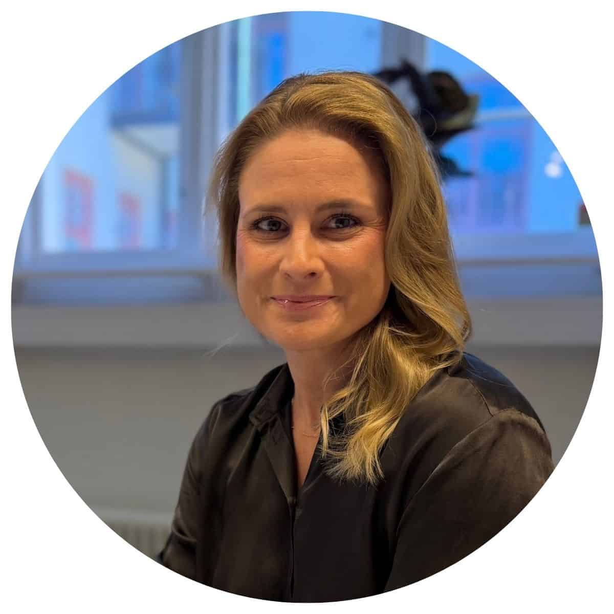 Kvinna med blont hår i svart blus, kontorsmiljö i bakgrunden.
